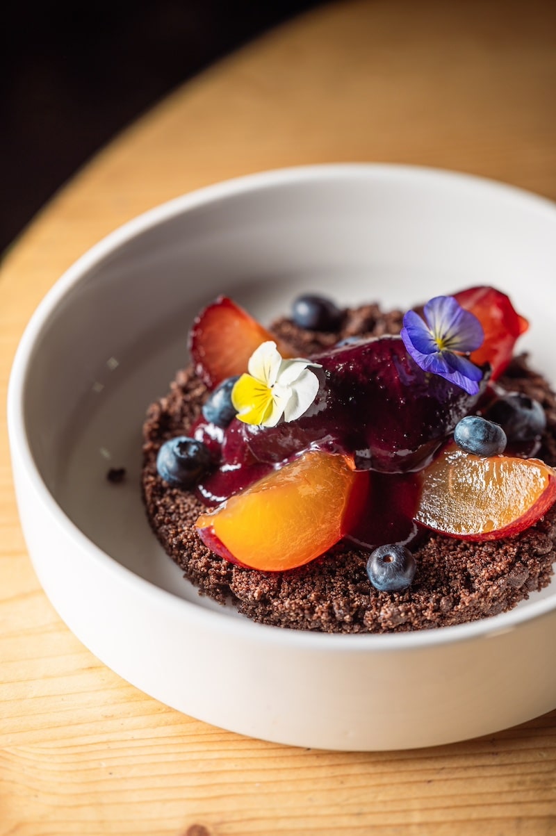 Dessert frais au chocolat, compote de fruits rouges, pêches, myrtilles et fleurs comestibles dans un bol blanc sur bois.