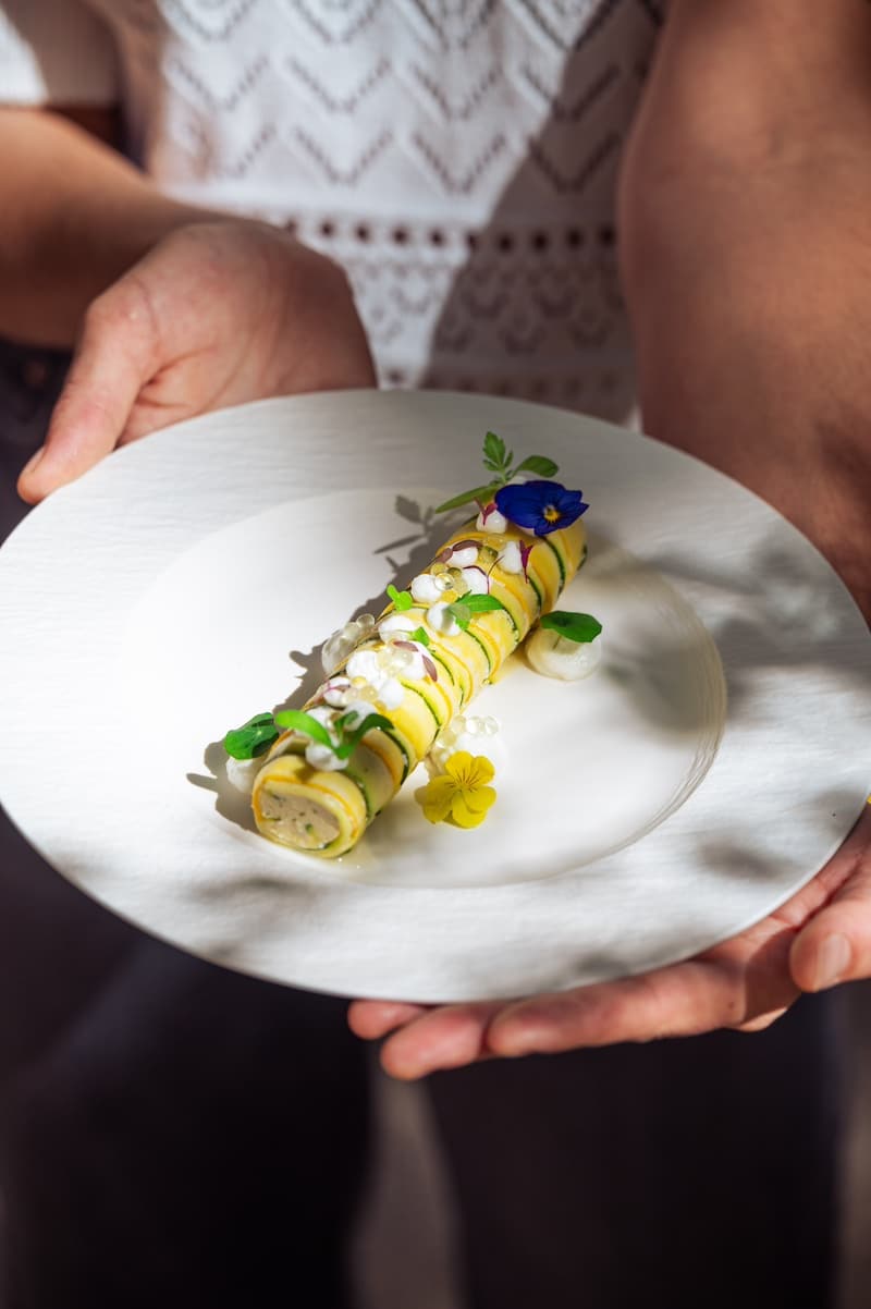 Personne tenant une assiette blanche avec un rouleau de courgette garni de caviar, crème et fleurs comestibles. Plat élégant.