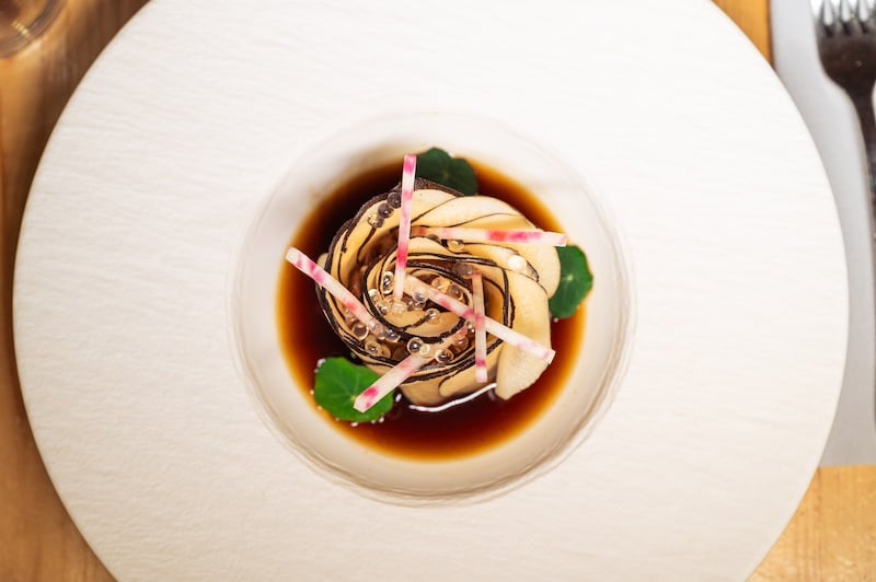 Plat gastronomique: spirale de légumes, perles et bâtonnets de radis roses dans une sauce brune, sur assiette blanche.