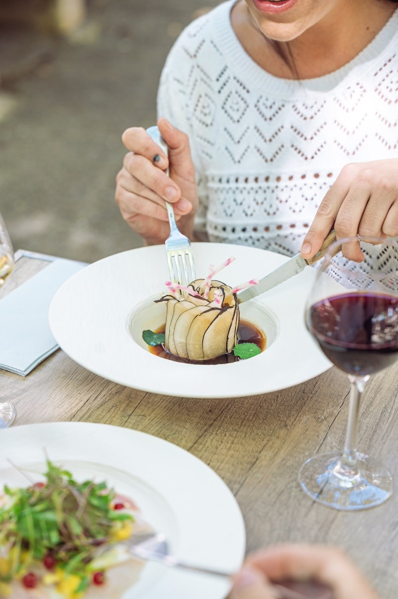 Femme dégustant un plat végétal raffiné en forme de fleur avec couteau et fourchette, verre de vin rouge à côté, repas élégant.