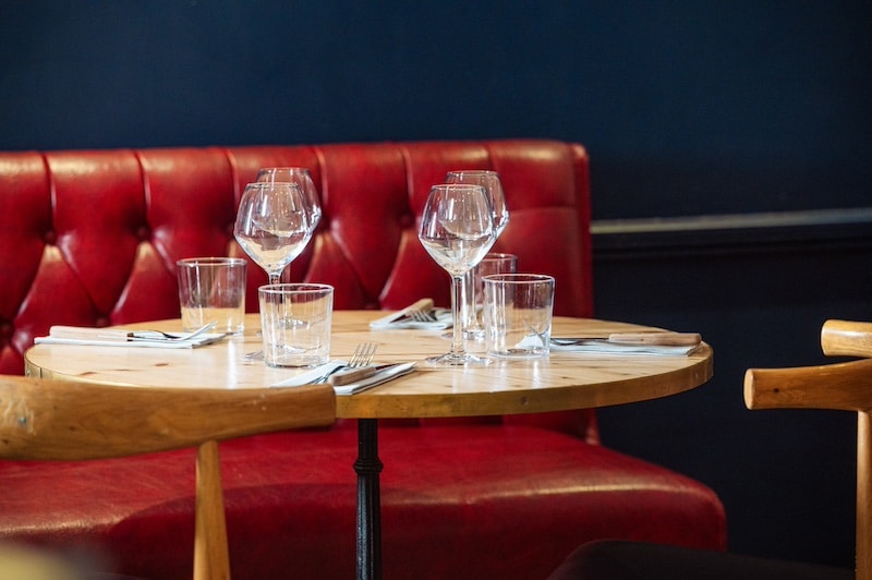 Table ronde en bois dressée avec verres et couverts devant une banquette capitonnée rouge vif dans un restaurant.