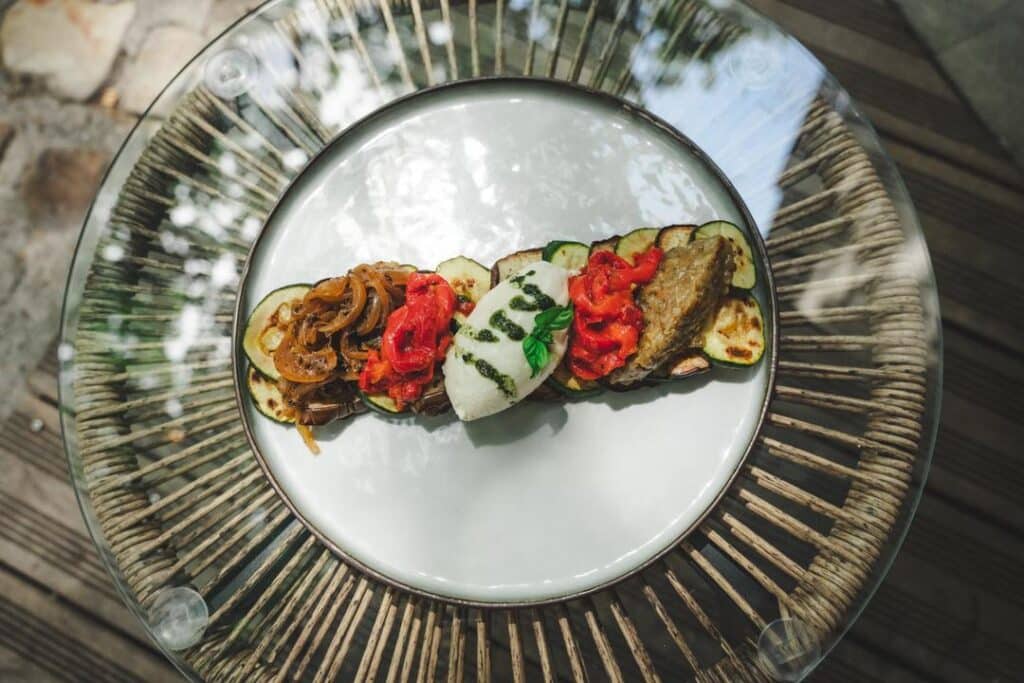 Plat gourmet d'été sur table en verre et rotin. Contient courgettes grillées, oignons caramélisés, poivrons rôtis, quenelle de fromage frais et pesto.