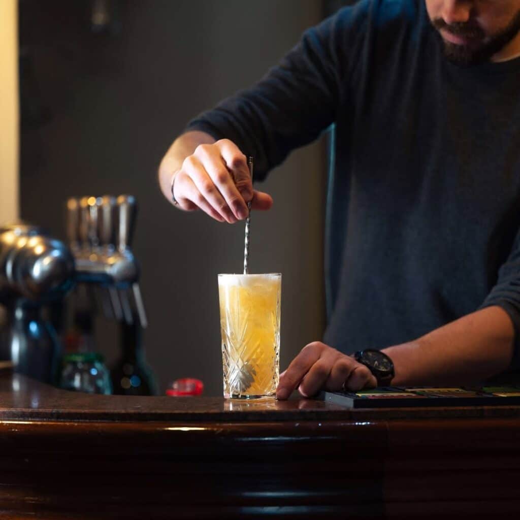 Barman prépare un cocktail jaune dans un verre texturé, remuant avec une longue cuillère. Scène de bar sombre et élégante.