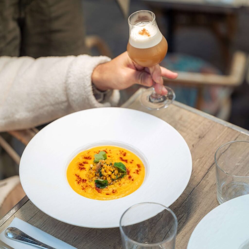Main tient bière artisanale mousseuse au-dessus d'un bol de soupe veloutée orange garnie. Table en bois, scène de restaurant.