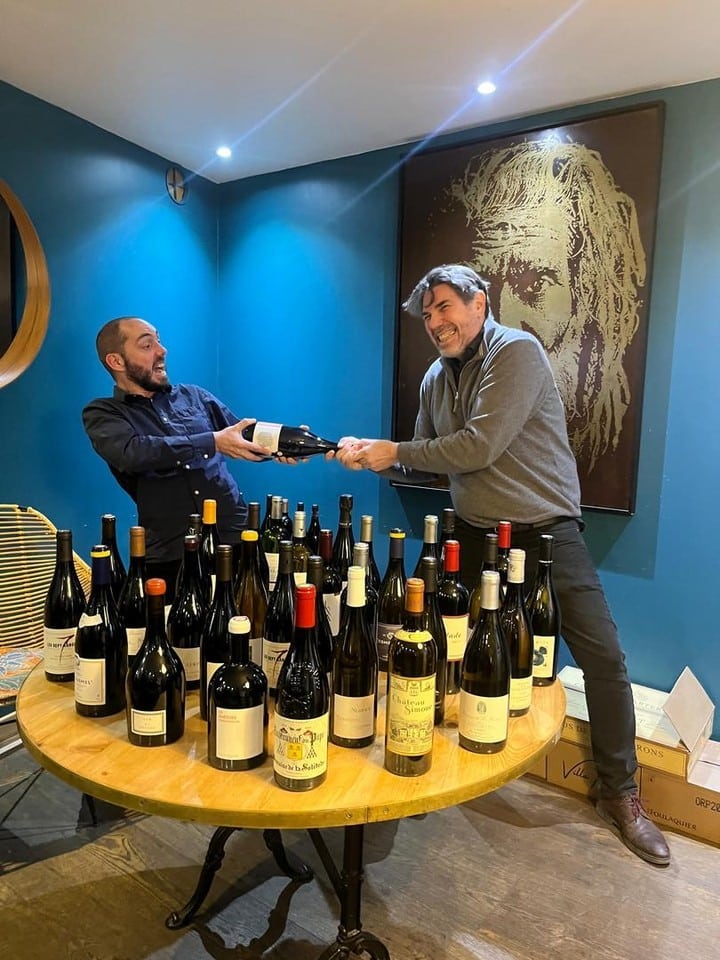 Deux hommes rieurs se disputent une bouteille de vin au milieu d'une table pleine de bouteilles. Ambiance conviviale.