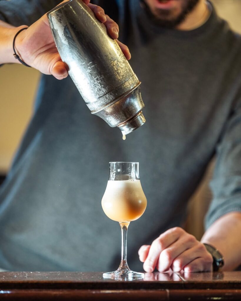 Homme en t-shirt gris versant un cocktail mousseux couleur pêche d'un shaker métallique dans un verre à pied. Une goutte pend du bec.