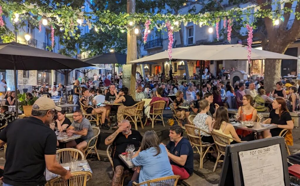 Terrasse de restaurant TÔT OU TARD animée en soirée. Des convives dînent sous des guirlandes lumineuses et parasols.