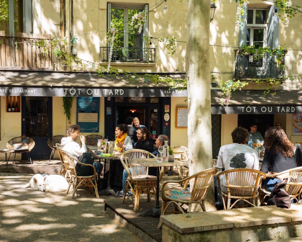 Clientèle détendue attablée en terrasse ensoleillée du café-restaurant TÔT OU TARD. Un chien blanc se repose au sol. Façade verdoyante.