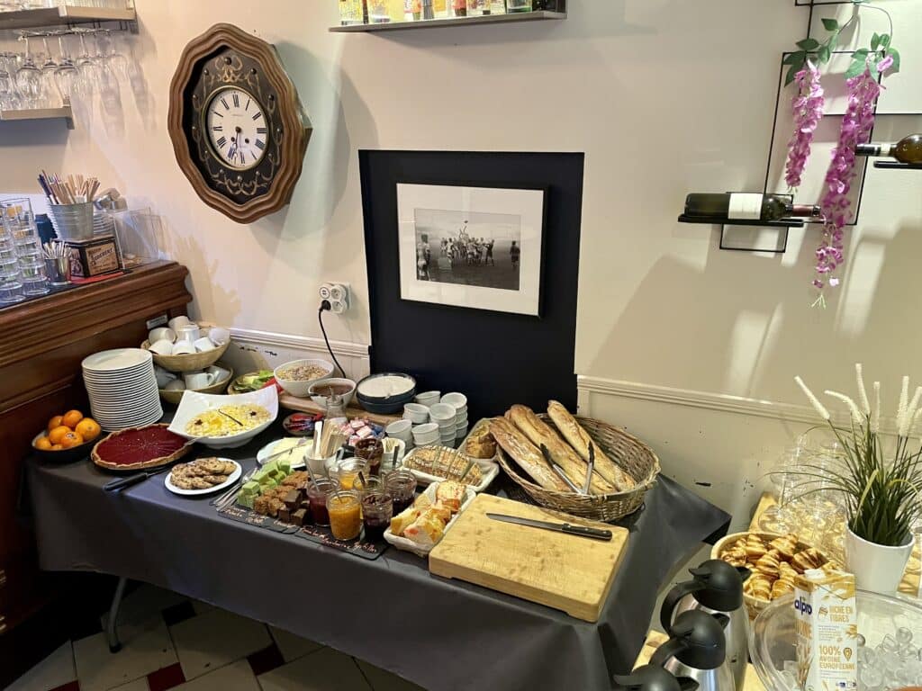 Buffet de petit-déjeuner continental avec assiettes, fruits, tartes, confitures, pain, céréales, yaourt et horloge murale ancienne.